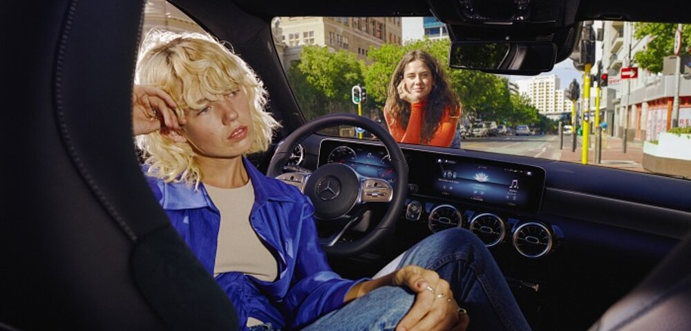Innenraum der A-Klasse Limousine mit Frau hinterm Lenkrad und einer Frau vor dem Auto, lehnend auf der Motorhaube